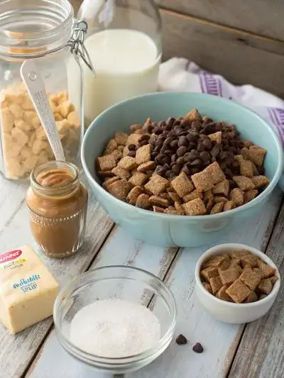Ingredients for puppy chow displayed on a kitchen table, including Rice Chex cereal, chocolate chips, peanut butter, butter, vanilla extract, and powdered sugar.