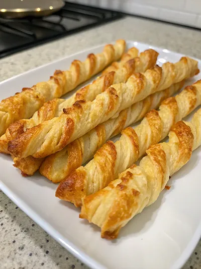 Golden puff pastry cheese twists stacked on a white plate.